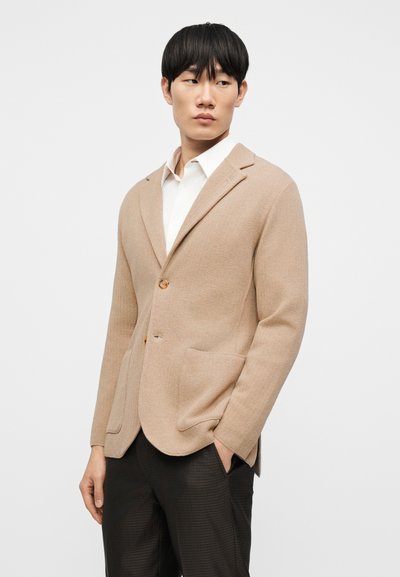 Μπεζ σακάκι με υφή, μονό κουμπί, γιακάδες με σχισμή και δύο εμπρόσθιες τσέπες, συνδυασμένο με λευκό πουκάμισο και σκουρόχρωμο παντελόνι με σχέδια.