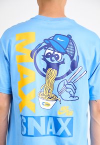Light blue cotton t-shirt featuring a cartoon character with headphones, holding noodles, and text "MAX IS SNAX" in yellow and blue.