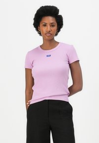 Lichtpaarse fitted t-shirt van ribbelstof. Beschikt over een ronde halslijn en een klein blauw merklabel aan de voorkant. Gecombineerd met zwarte broek.