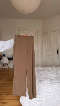 Persona sosteniendo pantalones anchos color beige en una percha en un dormitorio minimalista con suelo de madera, puertas blancas y ropa de cama blanca.