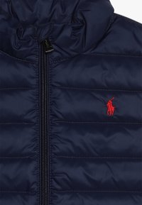 Chaqueta acolchada de navy con cierre de cremallera, que presenta un diseño acolchado y un logotipo bordado en rojo en el pecho. Textura suave y brillante.