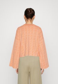 Cropped, textured cardigan in orange with wide sleeves. Paired with light green high-waisted pants, viewed from the back.