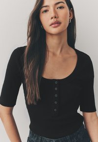 Top corto nero a costine con scollatura ampia a girocollo e chiusura frontale con sei bottoni neri. Tessuto liscio, design aderente.