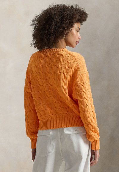 Πορτοκαλί πουλόβερ με πλεκτές πτυχώσεις, με ριπ τελείωμα και στρογγυλή λαιμόκοψη. Το σχέδιο διαθέτει υφασμένους μοτίβους και άνετη εφαρμογή, φορεμένο με ανοιχτόχρωμο παντελόνι.