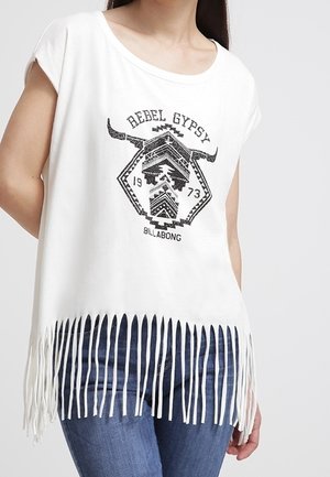 Kobieta ubrana w biały, bezrękawnik z frędzlami z napisem "Rebel Gypsy" i grafiką byka, w niebieskich dżinsach na jednolitym tle.