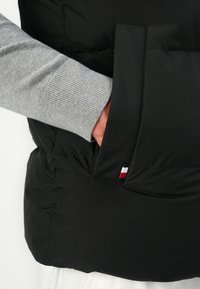 Hånd i lommen på en sort polstret vest, der bæres over en grå strikket ærme med et lille rød, hvid og blåt stofmærke på lommens kant.