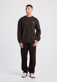 Felpa marrone con vestibilità rilassata, polsini e orlo a coste. Abbinata a pantaloni in velluto a coste marrone scuro e sneakers bianche con accenti verdi.