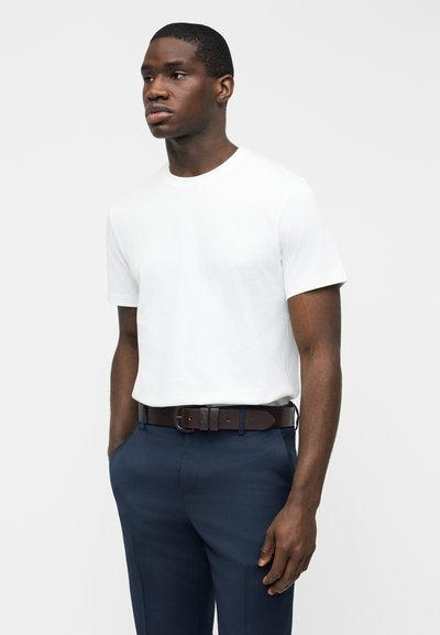 Άνδρας που φοράει απλό λευκό κοντομάνικο μπλουζάκι, σκούρο μπλε παντελόνι και σκούρο καφέ ζώνη, στέκεται με τα χέρια στις τσέπες.