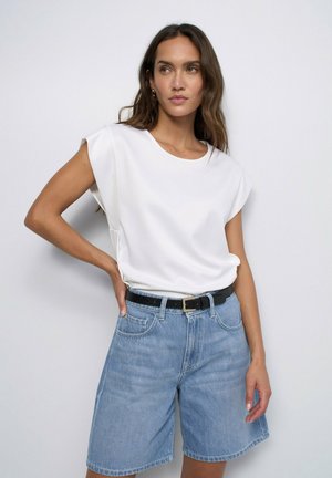 Femme aux longs cheveux bruns portant un haut blanc sans manches et un short en jean bleu clair, debout avec une main sur la taille devant un fond uni.