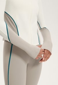 Jachetă sport cu mâneci lungi, cu fermoar, de culoare albă, cu accente teal, asortată cu colanți bej ajustați. Textură netedă și design minimalist.
