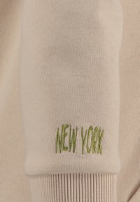 Felpa a coste beige con ricamo verde "NEW YORK" sul polsino. Texture morbida con vestibilità aderente.