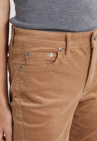 Braune Cordhosen mit strukturierter Oberfläche, die über Vorderaschen, Metallnieten und einen hohen Bund verfügen. Der Stoff ist weich und gerippt.