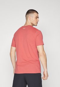 Rød t-skjorte med korte ermer i et mykt, glatt stoff med rund hals og liten logo bak. Båret med mørke shorts, sidevisning.