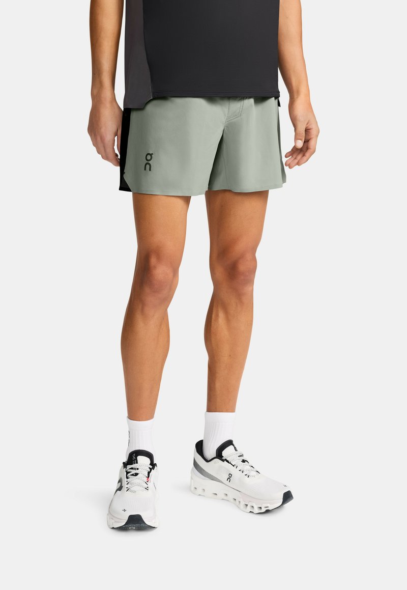 Persona con pantalones cortos de running verde claro, zapatos deportivos blancos con detalles negros, calcetines blancos y una camiseta negra, todo sobre un fondo blanco.