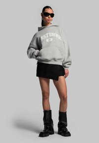 Felpa grigia con cappuccio e testo "RSTBHVR", gonna mini nera e stivali al ginocchio neri. La modella indossa occhiali da sole neri e mostra uno stile casual.