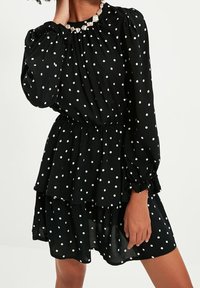 Abito nero con pois, dotato di lunghe maniche a sbuffo, vita elastica e gonna a strati con balze. Il tessuto appare leggero e strutturato.