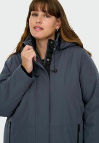 Sieviete ar gaiši brūniem matiem, valkājot tumši pelēku kapuci ar jaku, tur plecu apkakli pie sejas pret vienkāršu gaišu fonu.