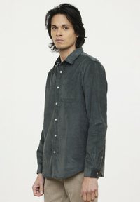 Chemise en velours côtelé vert foncé avec un devant à boutons, un col et une poche poitrine unique. Manches longues avec poignets boutonnés et tissu texturé.