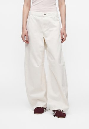 Personne portant un pantalon blanc ample à jambes larges et des baskets rouge foncé, debout devant un fond blanc uni.