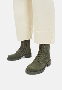 Tumši zaļi zamša kaujas zābaki ar masīlu zoli, valk�āti ar krēmkrāsas platām biksēm, kas ir nogrieztas virs potītēm.