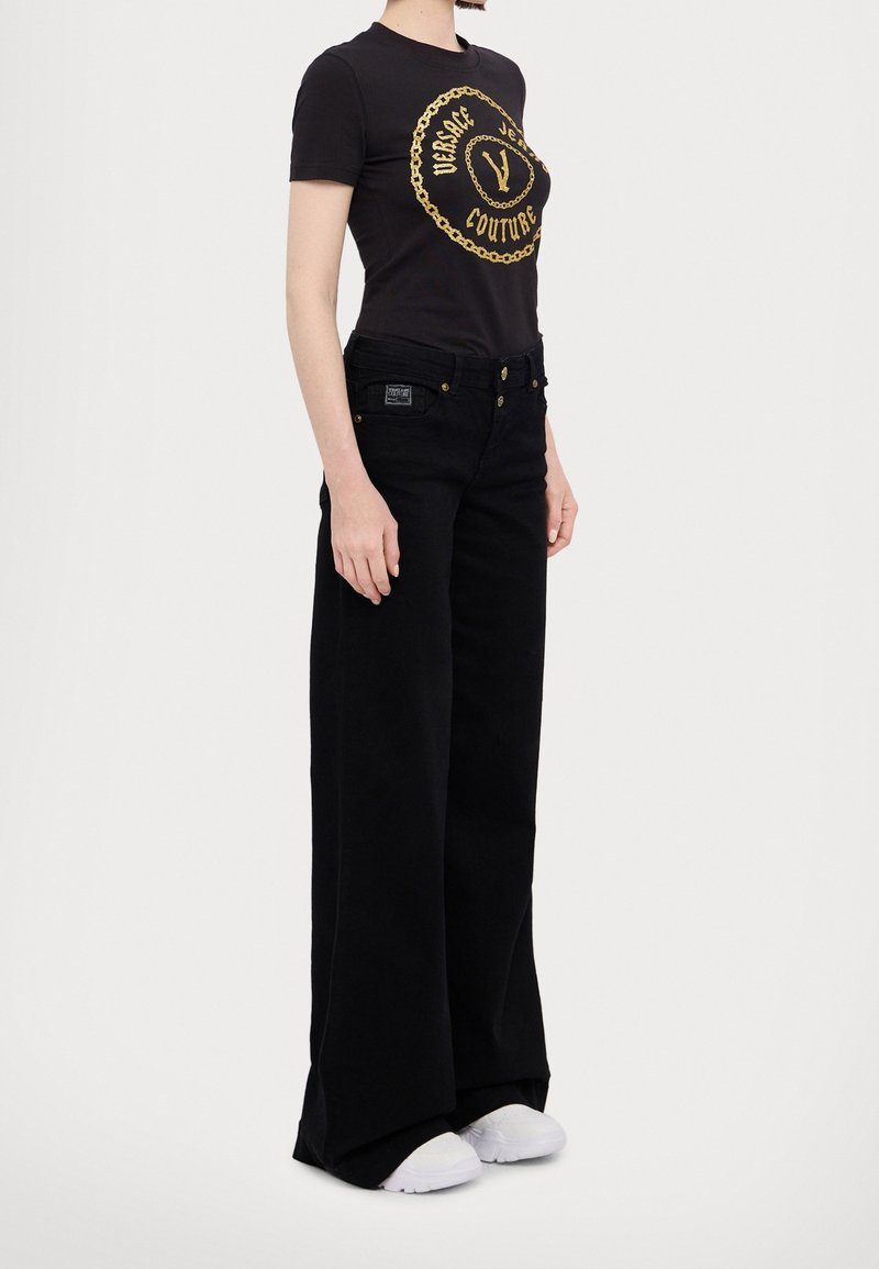 Femme portant un t-shirt noir Versace Jeans Couture avec logo doré, pantalon noir à jambes larges et baskets blanches, debout devant un fond blanc.