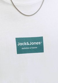 Άσπρο πουκάμισο με λογότυπο Jack & Jones σε τυρκουάζ και το σύνθημα "Definition of Denim", συνοδευόμενο από ασημένιο κολιέ αλυσίδα.