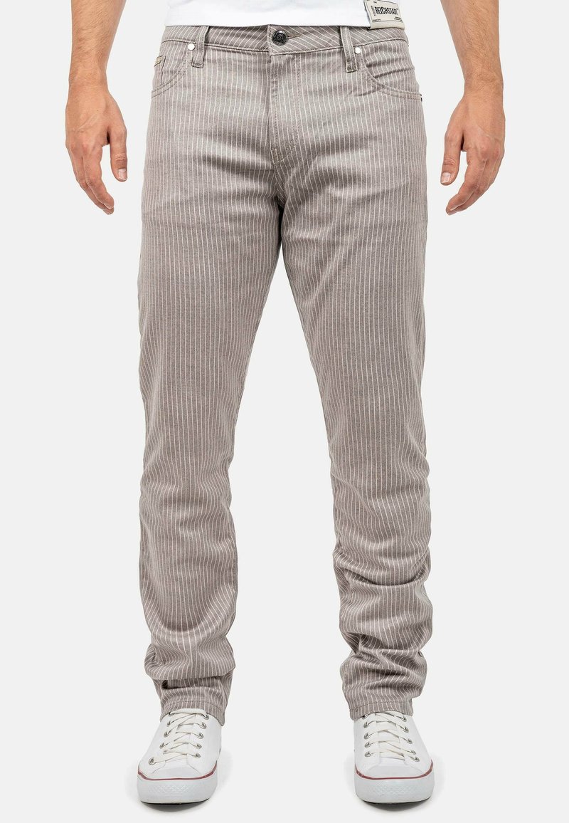 Homme portant un pantalon beige à fines rayures et des baskets blanches, debout les mains le long du corps devant un fond uni.