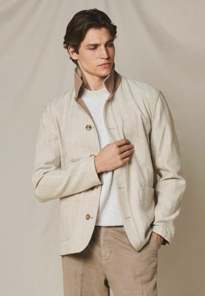 Mann, der eine beige Jacke mit Knöpfen und hochgestelltem Kragen anpasst, trägt ein weißes Hemd und hellbraune Hose, vor neutralem, einfarbigem Hintergrund stehend.