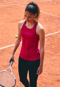 Svetlo roza športna majica brez rokavov z črnimi poudarki, v kombinaciji s črnimi legicami. Manekenka drži belo teniško lopar na peščeni teniški igri.