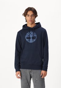 Navyblauwe hoodie, zachte stof, met een groot lichtblauw boompje-logo op de borst, kangoeroezak en rib gebreide manchetten en onderrand.