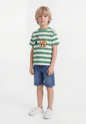 Blond gutt står og har på seg grønn og hvit stripete t-skjorte med oransje krantrykk, blå denimshorts og hvite joggesko.