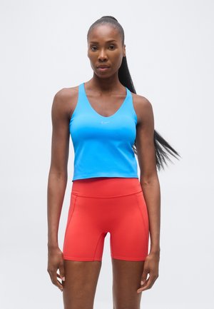 Športovo vyzerajúca žena s dlhým copom, oblečená v jasne modrom tielku bez rukávov a červených športových krátkych nohaviciach s vysokým pásom, stojaca pred jednoduchým svetlým pozadím.