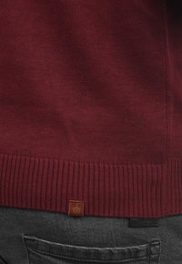 Burgunderfarbener Strickpullover mit geripptem Saum, versehen mit einem Logotag. Weiche Textur und taillierte Passform, mit einem schlichten, klassischen Erscheinungsbild.