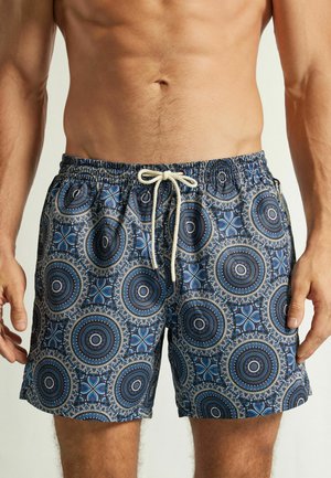Männlicher Oberkörper mit blauen Badeshorts mit kreisförmigen Mandalamustern und weißem Kordelzug, der in der Taille gebunden ist.