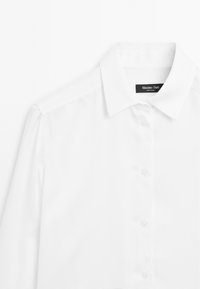Camicia bianca con colletto strutturato, realizzata in tessuto liscio. Presenta una patta frontale con bottoni trasparenti e un'etichetta con logo all'interno.