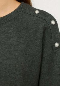 Pullover grigio scuro con scollo a girocollo, caratterizzato da due bottoni decorativi con dettagli in perla sulla spalla, realizzato in materiale morbido a maglia.