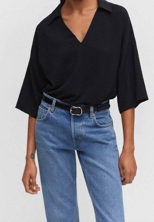Frau trägt ein schwarzes V-Ausschnitt-Oberteil mit weiten Ärmeln, das in eine hoch taillierte blaue Jeans mit einem dünnen schwarzen Gürtel mit goldener Schnalle gesteckt ist.
