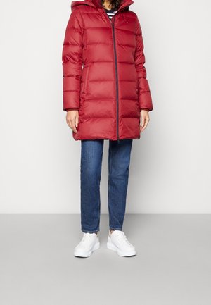 Personne portant un manteau matelassé rouge au genou avec une capuche, un jean bleu et des baskets blanches, debout devant un fond uni.