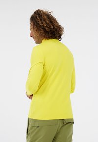 Man met krullend haar in een heldergele lange mouwen shirt en olijfgroene broek, iets naar de zijkant gekeerd tegen een witte achtergrond.