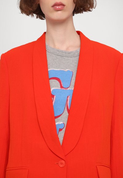 Bright orange blazer with a shawl collar, single button closure, and a textured finish, layered over a gray t-shirt with blue graphic print.