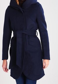 Marinblå fleecejacka med huva, bältad midja och framfickor. Har rundade sömmar och en scalloped nederkant för extra detalj.