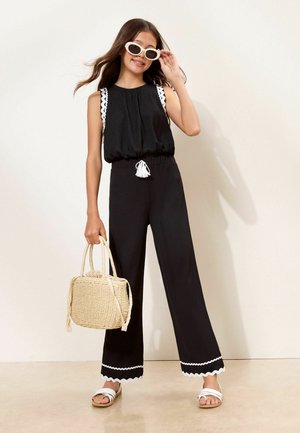 Junge Frau in schwarzem ärmellosem Oberteil und Hose mit weißem Besatz, trägt weiße Sonnenbrille und Sandalen, hält eine beige geflochtene Handtasche.