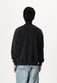 Uomo che indossa un ampio maglione nero e jeans azzurri chiari, in piedi con le spalle alla macchina fotografica su uno sfondo bianco semplice.