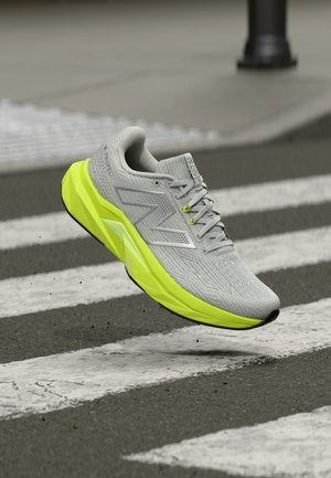 Scarpa da ginnastica grigia e giallo neon in aria sopra una striscia pedonale su asfalto, che mostra il profilo laterale con il logo del marchio visibile e suola testurizzata.