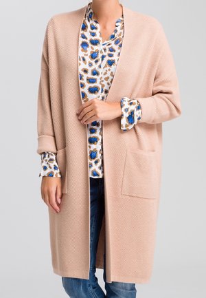 Femme portant un long cardigan en maille beige sur une blouse blanche à imprimé léopard bleu et marron, assorti à un jean bleu.