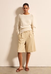 Cremfarbener Pullover mit strukturiertem Material und geripptem Saum, beige Faltenkurze, und braunen geflochtenen Sandalen. Einfache, lässige Formen mit hellen Farben und glatten Texturen.