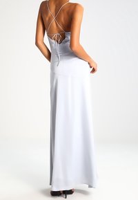 Kvinna iklädd en golvlång vit klänning med korsande ryggband, stående med ena handen vilande vid höften.