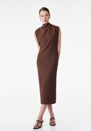 Kobieta ubrana w brązową, bezrękawną midi sukienkę z wysokim kołnierzem oraz czarne sandały na szpilce, stojąca na tle jednolitej białej ściany.