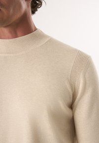 Beige gebreide trui met een gladde textuur en ribbelige ronde hals. Heeft textuuraccenten op de schouders en een getailleerd silhouet.