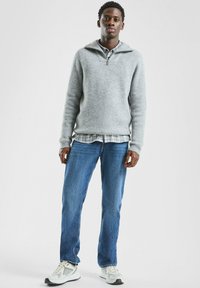 Szary dzianinowy sweter z kołnierzem na ćwiartkę zamka, noszony na koszuli w kratę, zestawiony z niebieskimi dżinsami i szarymi sneakersami z białymi akcentami.